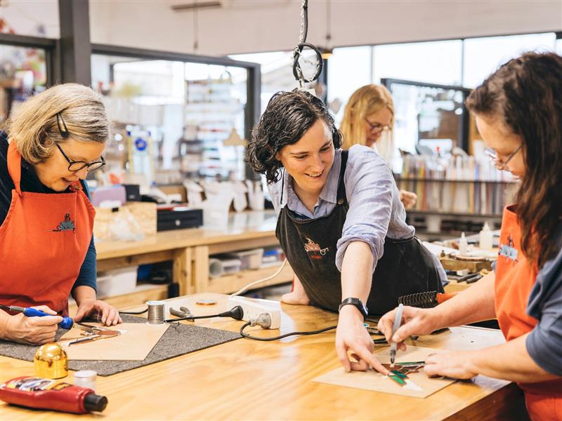 two women in red aprons working on stained glass with a teacher in a black apron helping