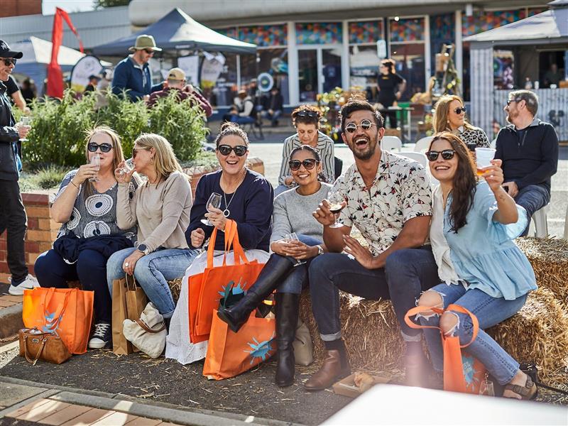 A photo of six people sitting on hay bales wearing sunglasses, smiling, holding glasses of wine