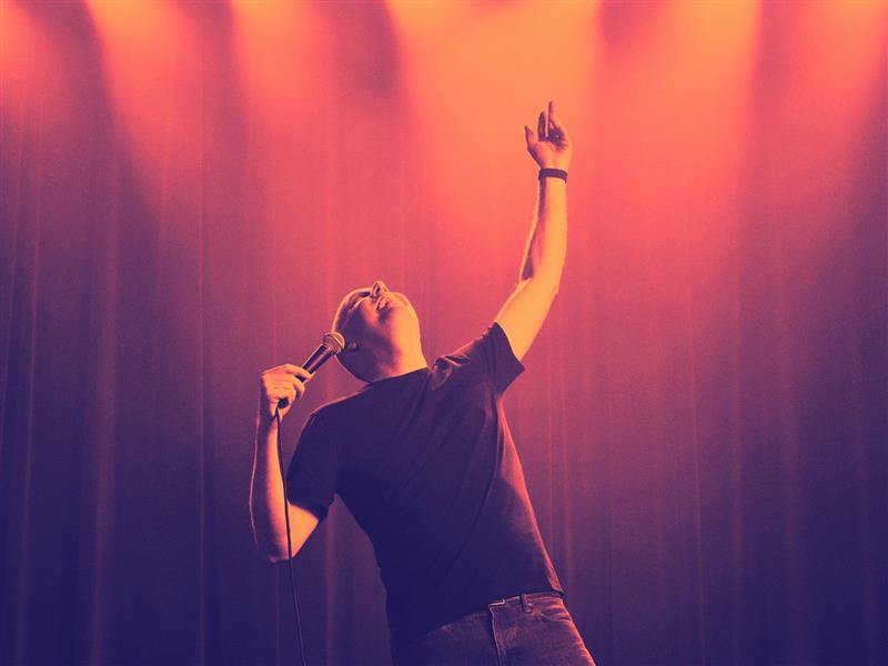 Tom on stage reaches toward the sky while holding microphone
